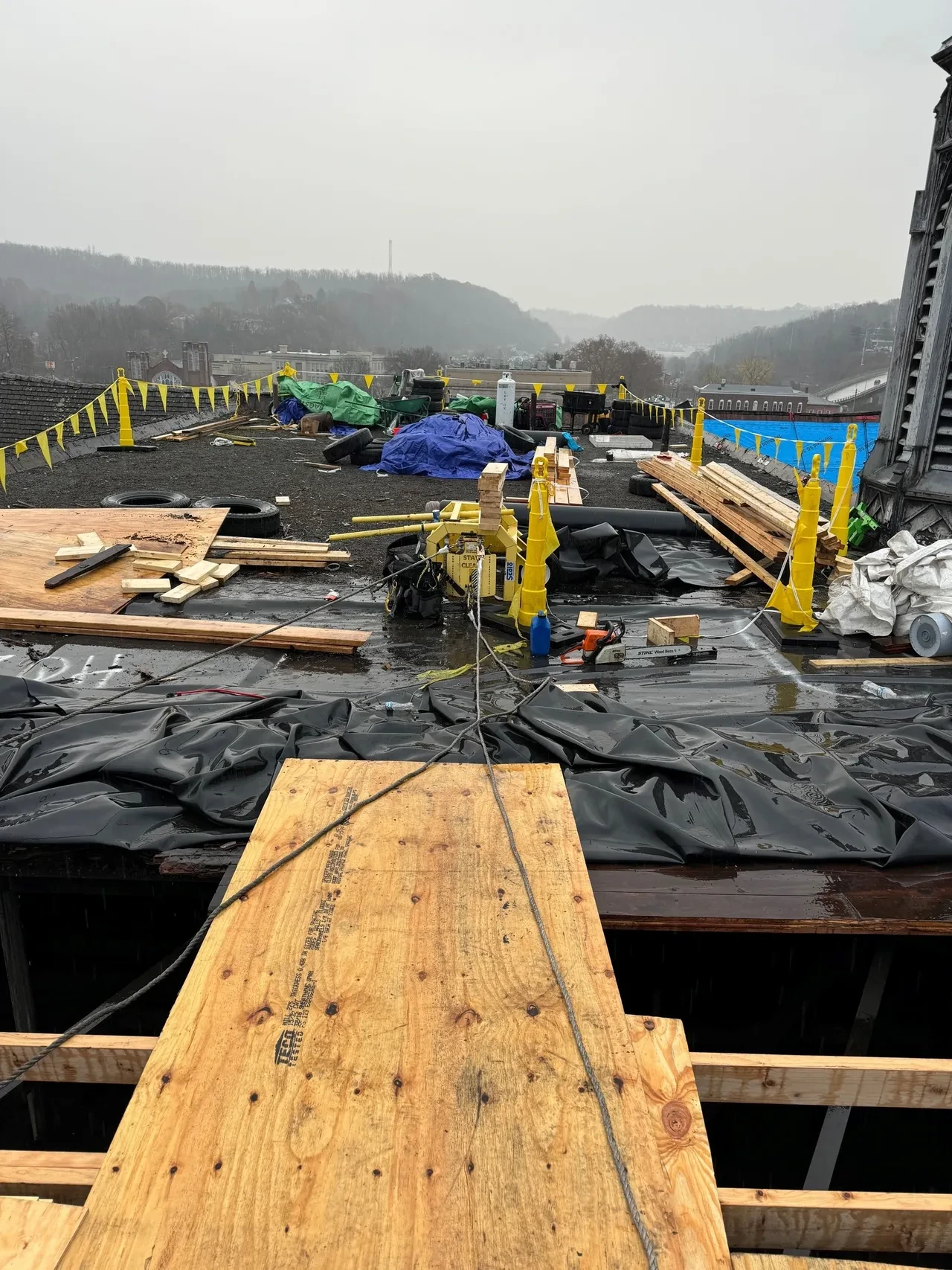 Worker in hi-vis vest and harness laying new roof decking with Pittsburgh skyline visible in background